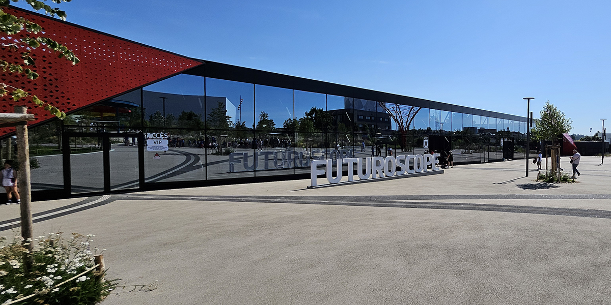 L'entrée principale du parc du Futuroscope se distingue par une façade vitrée moderne et épurée, reflétant l’extérieur et surplombée d’une grande enseigne blanche marquant le nom emblématique du lieu.