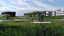 Des équipements de fitness en plein air sont installés au bord d’un plan d’eau, au cœur d’un espace vert entouré de bâtiments modernes, dans un cadre paisible et propice à l’activité physique.