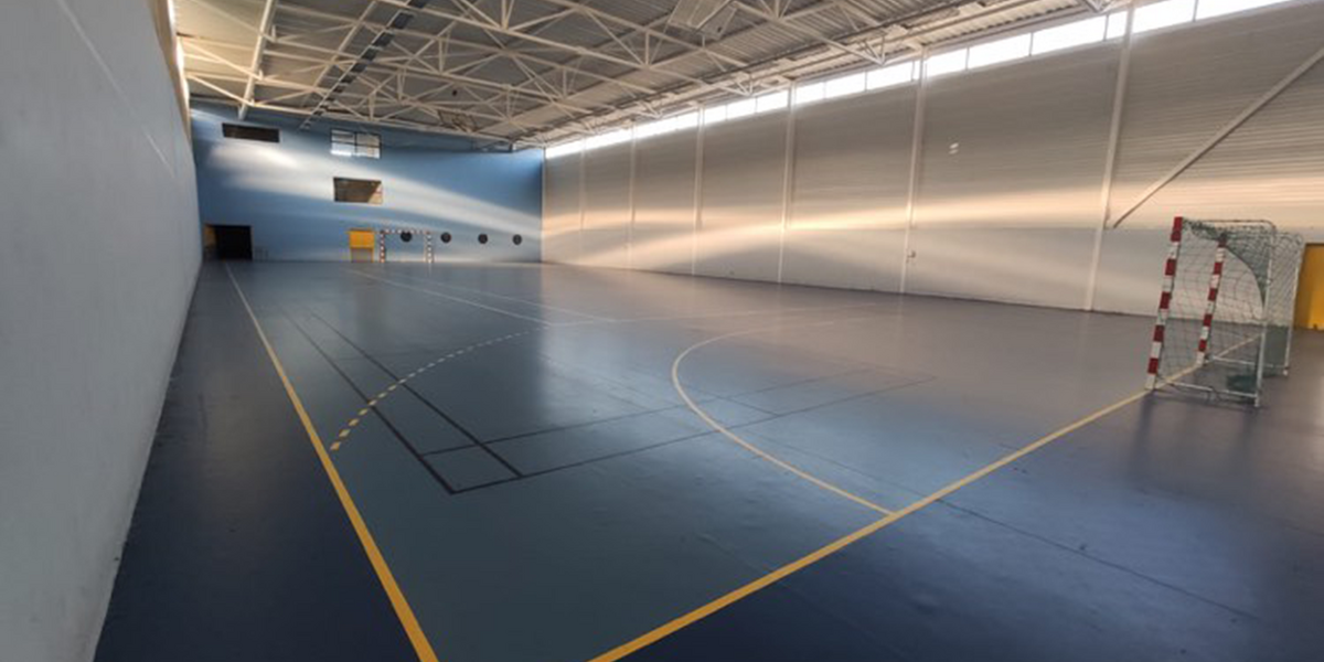 Une grande salle de sport intérieure, éclairée par la lumière naturelle, avec des marquages au sol pour plusieurs disciplines et des buts de handball placés à chaque extrémité.