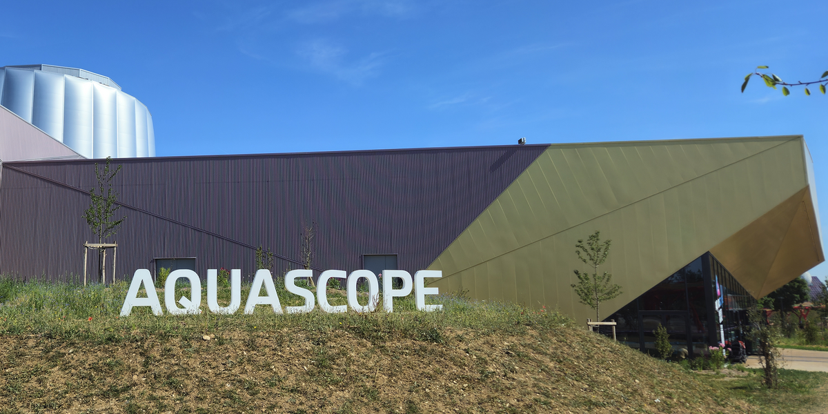 Le bâtiment au design géométrique et coloré porte le nom d’"AQUASCOPE", suggérant un lieu dédié à l’eau ou à l’exploration scientifique, implanté dans un environnement moderne et végétalisé.
