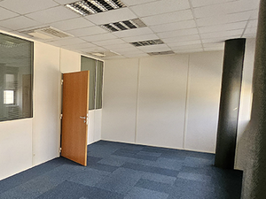 Un bureau vide à la moquette bleue et aux cloisons blanches, doté de fenêtres intérieures vitrées, d’une porte en bois et d'une colonne sombre qui contraste avec l’espace clair.