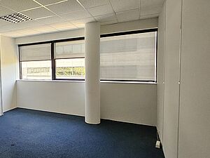 Un bureau vide, sobre et lumineux, avec moquette bleue, murs blancs, grandes fenêtres partiellement occultées et une colonne blanche en son centre.